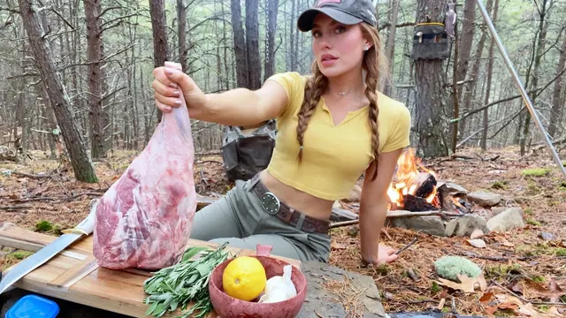 Heidi holding up a leg of lamb beside a campfire in pine woods